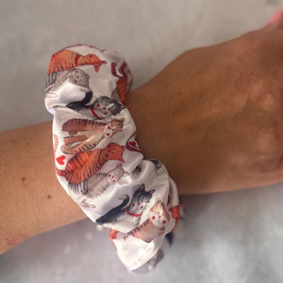 A close-up of a fabric hair scrunchie worn on a person's wrist. The scrunchie has a white background with a printed pattern of various cats in orange, black, gray, and white, interspersed with small red hearts.