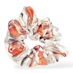 A satin scrunchie featuring an all-over print of orange, red, and grey tabby cats on a white background, shown against a plain white studio backdrop.
