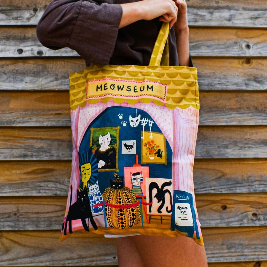 A person holds up a colorful tote bag with a whimsical cat-themed illustration and a yellow 'MEOWSEUM' sign