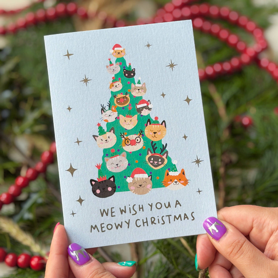 A person holds a light blue Christmas greeting card with an illustration of a cat-shaped Christmas tree and the text 'WE WISH YOU A MEOWY CHRISTMAS'. 