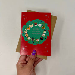 A hand holds a red Christmas greeting card featuring a green wreath with cartoon animal faces and the text 'Merry Midwing Christmas,'