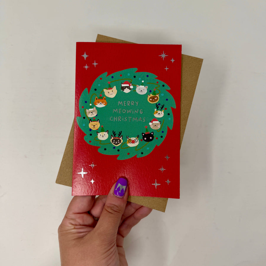 A hand holds a red Christmas greeting card featuring a green wreath with cartoon animal faces and the text 'Merry Midwing Christmas,'