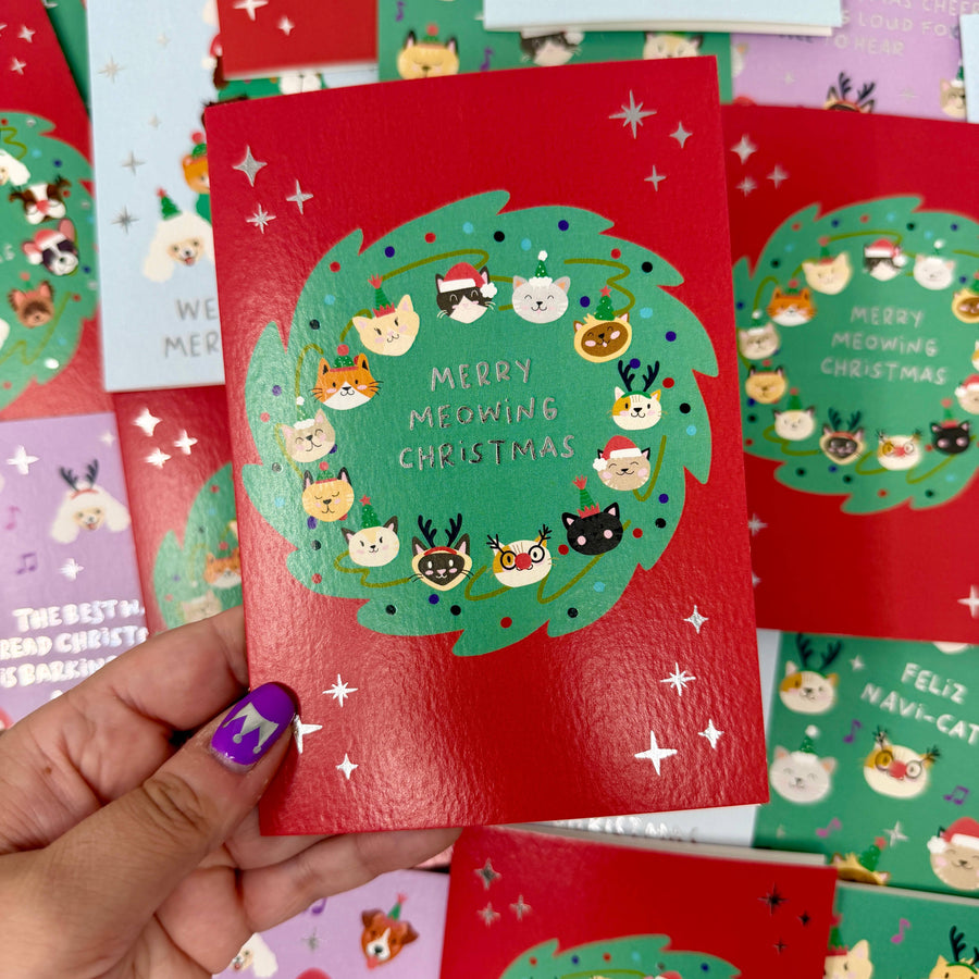 A hand holds a red Christmas greeting card featuring a green wreath with cute cat faces and the text 'MERRY MEOWING CHRISTMAS.'