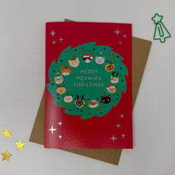 A red Christmas greeting card with a green wreath illustration and the text 'MERRY MEOWING CHRISTMAS' surrounded by small cat faces. 