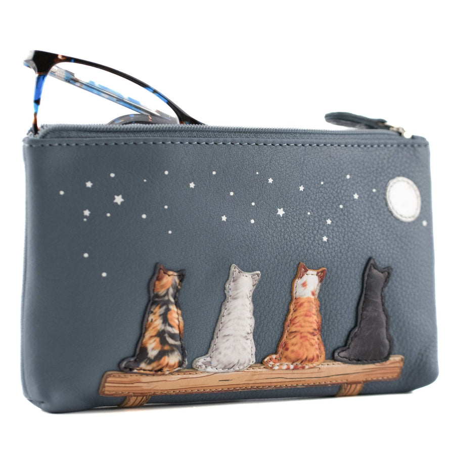 A slate blue pebbled leather zippered pouch, featuring an illustration of four cats sitting on a bench under a starry night sky. A pair of eyeglasses is tucked into the top zipper.