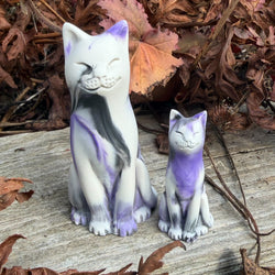 Two ceramic cat figurines, one large and one small, decorated with white, purple, and black marbled patterns, set outdoors on a weathered wooden surface surrounded by dry autumn leaves.