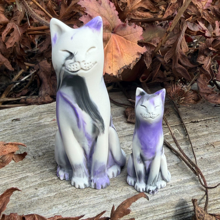 Two ceramic cat figurines, one large and one small, decorated with white, purple, and black marbled patterns, set outdoors on a weathered wooden surface surrounded by dry autumn leaves.