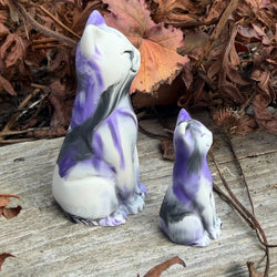 Two ceramic cat figurines, one large and one small, decorated with a white base and purple and black marbled patterns, placed on a weathered wooden surface with dry autumn leaves in the background.