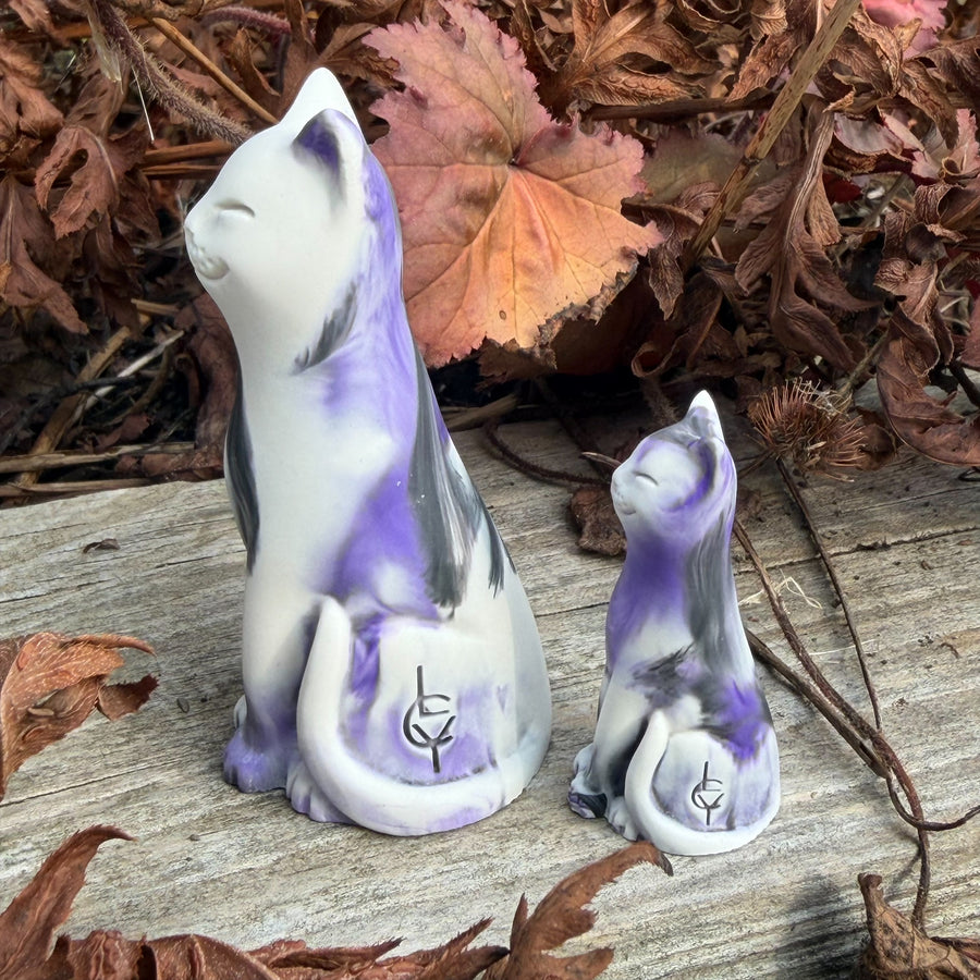 Two ceramic cat figurines, one large and one small, decorated with white, purple, and gray marbled patterns, displayed on a weathered wooden surface surrounded by dried autumn leaves.