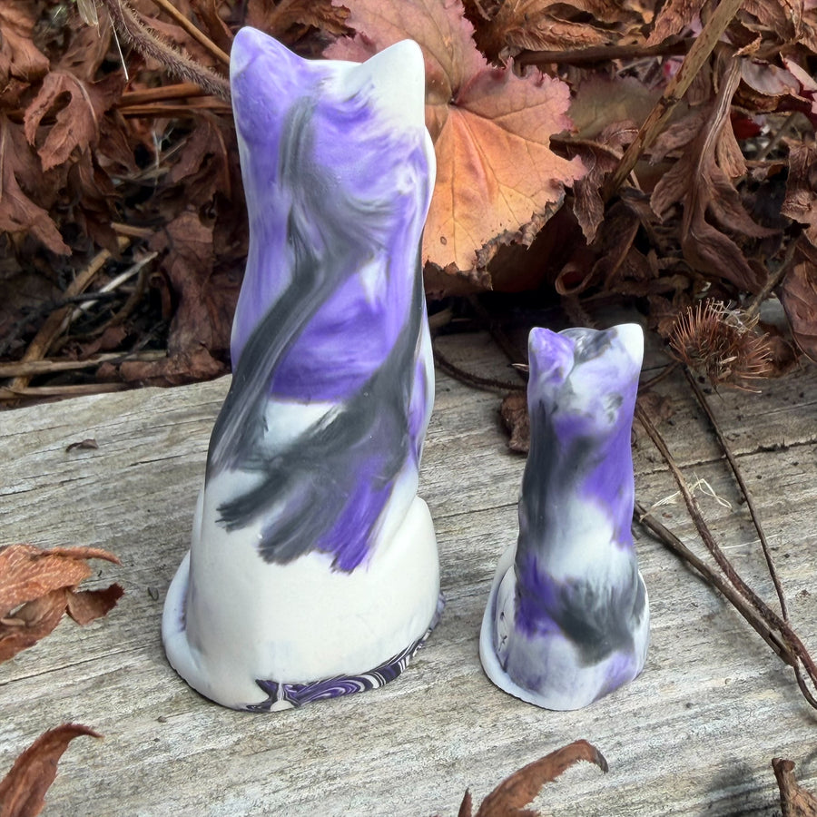 Two ceramic cat figurines, one large and one small, featuring a purple, white, and black marbled pattern, displayed outdoors on a weathered wooden surface with dry leaves in the background.