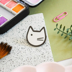 An enamel pin badge with a minimalist black and white illustration of a cat's face near paints