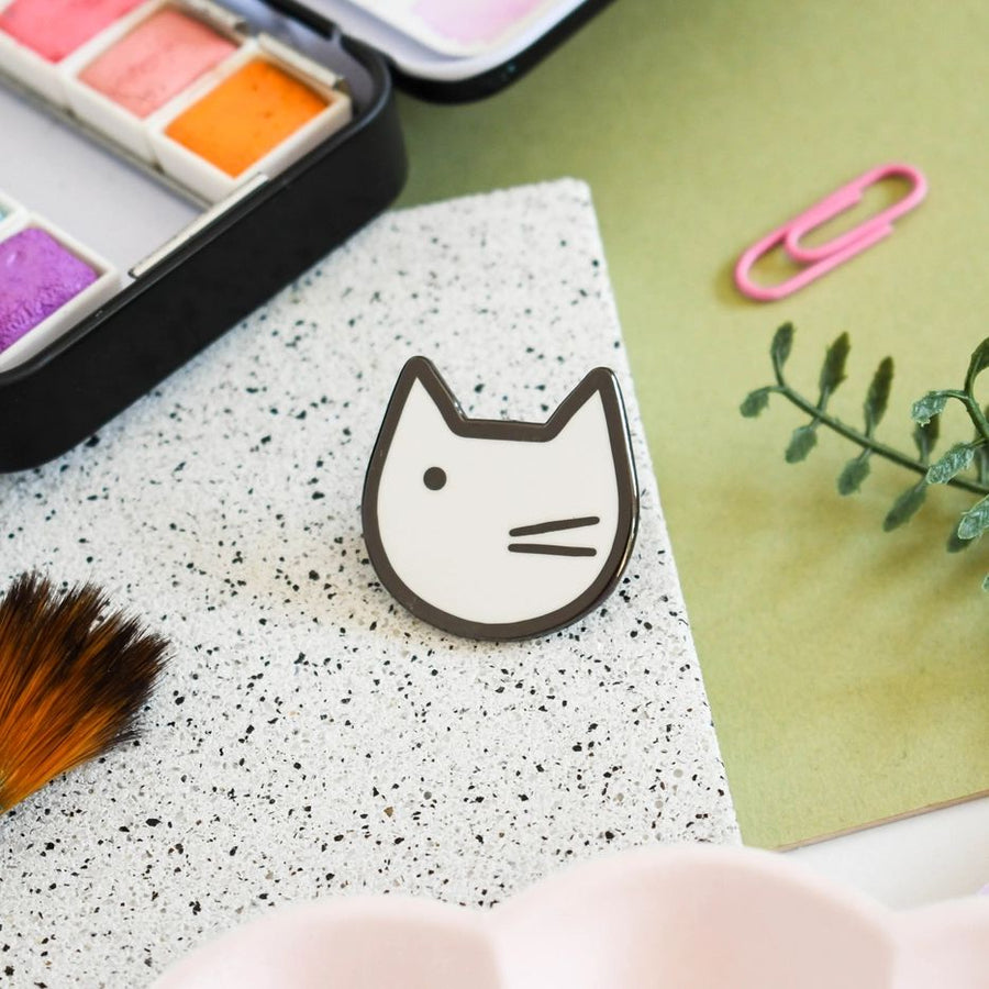 An enamel pin badge with a minimalist black and white illustration of a cat's face near paints