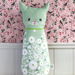 A mint green cat-shaped lumbar pillow with a white daisy floral pattern, sitting on a light beige surface against a pink and black bird-patterned wallpaper background.