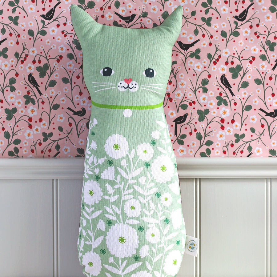 A mint green cat-shaped lumbar pillow with a white daisy floral pattern, sitting on a light beige surface against a pink and black bird-patterned wallpaper background.