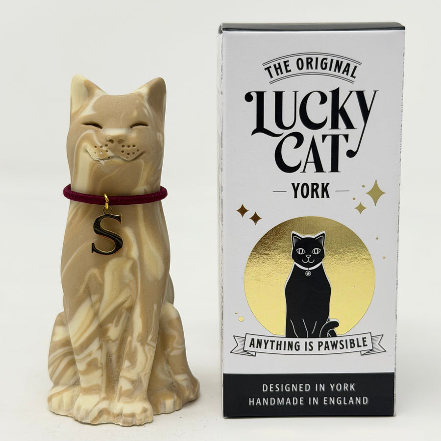 A creamy brown swirl resin lucky cat figurine wearing a burgundy collar with a black letter 'S' charm, standing next to its white, black, and gold retail box labeled 'The Original Lucky Cat York'.
