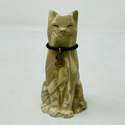 A resin lucky cat figurine in a sitting position, featuring a cream and tan marbled pattern, closed eyes, a small smile, and a dark red collar with a gold letter 'S' charm.