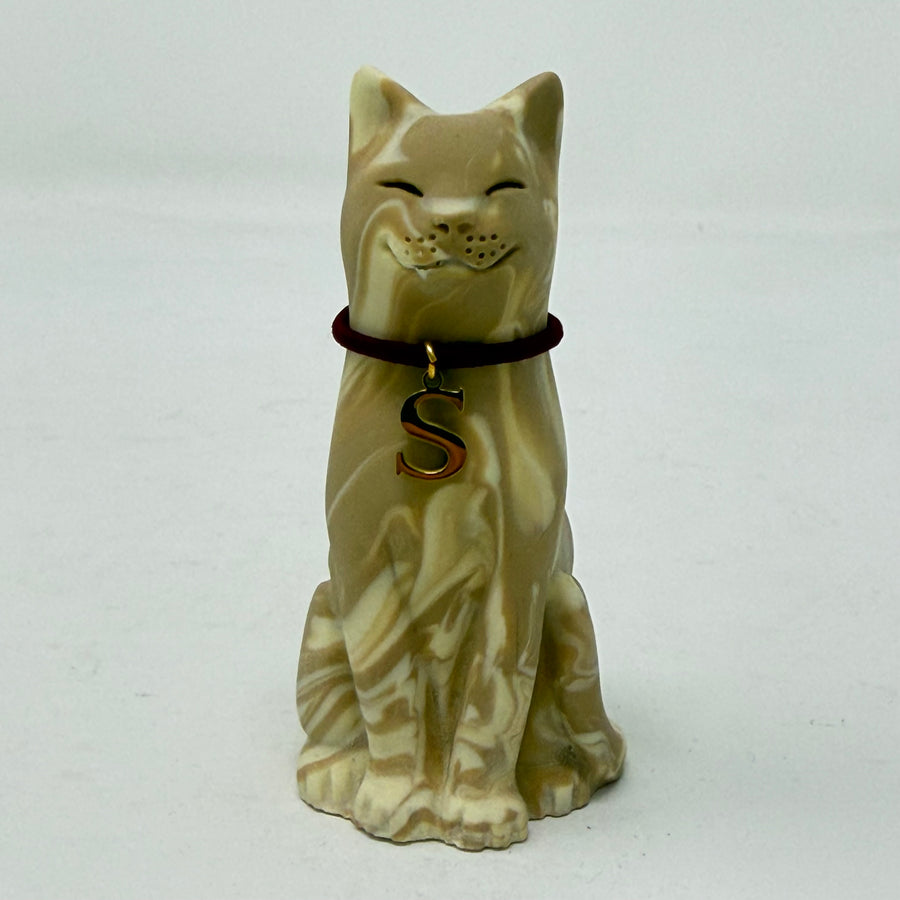 A resin lucky cat figurine in a sitting position, featuring a cream and tan marbled pattern, closed eyes, a small smile, and a dark red collar with a gold letter 'S' charm.
