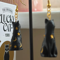 Black cat-shaped dangle earrings with gold stars and crescent moons, displayed on a black stand next to a Lucky Cat box.