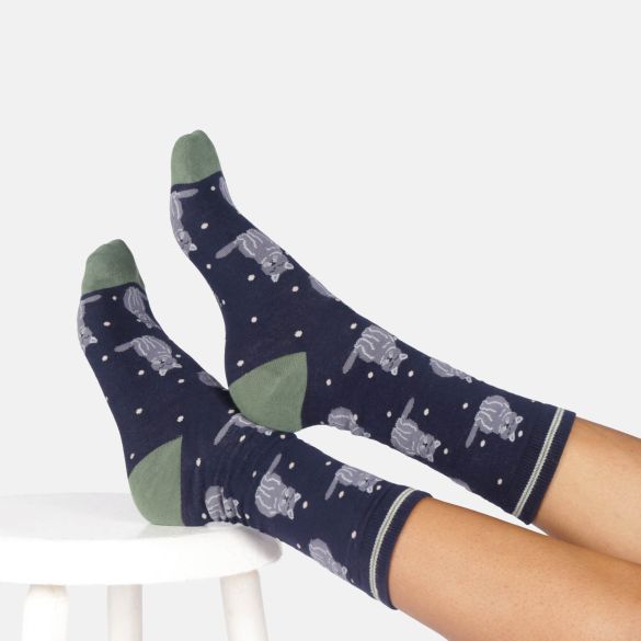 A pair of navy blue crew socks with a grey cat and white polka dot pattern, shown on a person's feet resting on a white stool.
