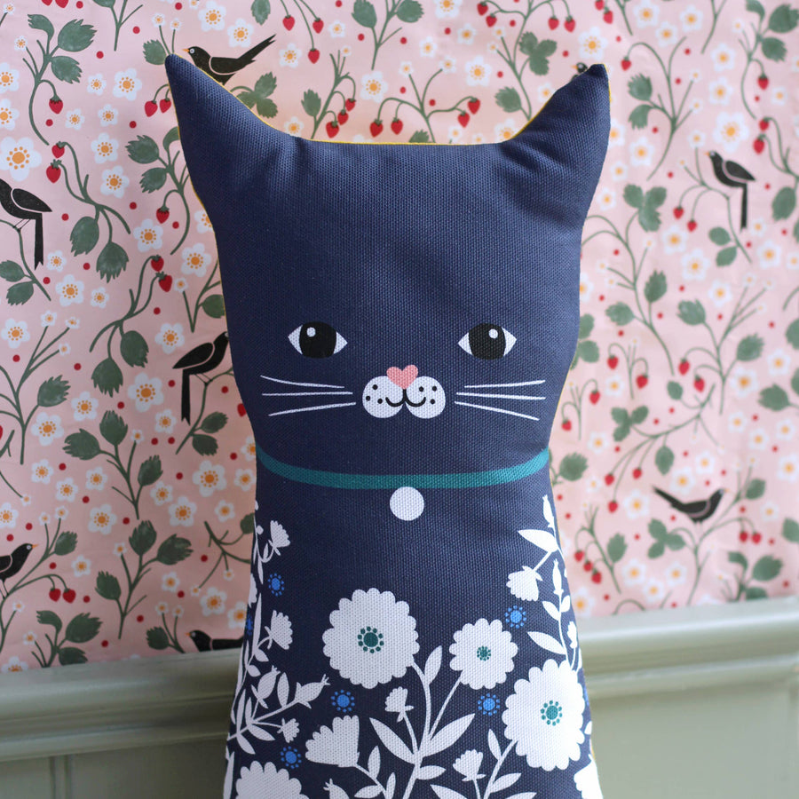 A navy blue cat-shaped decorative pillow with a floral patterned lower half, featuring embroidered black eyes, white whiskers, a pink nose, and a teal collar, set against a pink floral wallpaper background.
