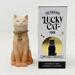An Apricot resin lucky cat figurine wearing a gold collar with a letter 'S' charm, standing next to its branded box, 'The Original Lucky Cat York'