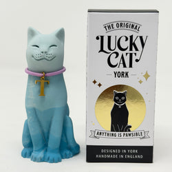 A light blue and turquoise lucky cat figurine wearing a pink collar with a gold cross pendant, shown next to its branded box labeled 'The Original Lucky Cat York'.