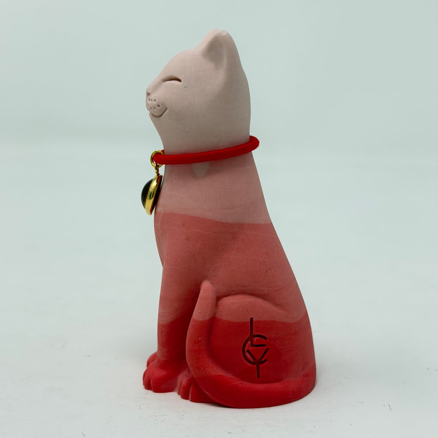 A resin lucky cat figurine in a seated position, in multi tone red colour scheme, wearing a red collar with a small gold charm