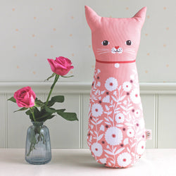 A pink cat-shaped decorative pillow with a white floral pattern, styled on a white surface next to a small glass vase with pink roses against a light gray paneled wall.