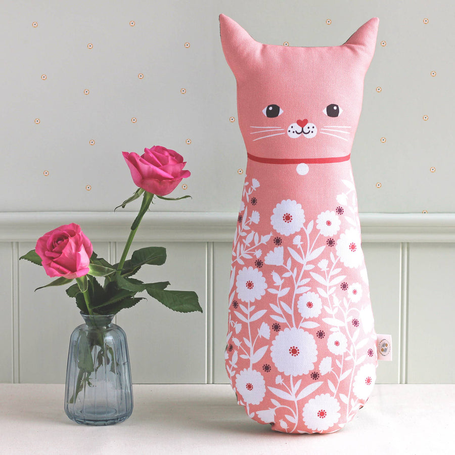 A pink cat-shaped decorative pillow with a white floral pattern, styled on a white surface next to a small glass vase with pink roses against a light gray paneled wall.