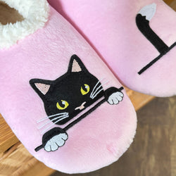 Pink plush slippers with a black cat face design and white faux-fur trim, shown on a wooden surface