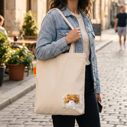 A woman carrying a beige canvas tote bag on a cobblestone street. The tote bag features a small, realistic plush ginger cat peeking out from a ruffled white fabric edge near the bottom right corner
