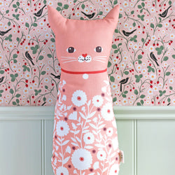A pink cat-shaped decorative pillow with a floral pattern, featuring a simple embroidered face, set against a floral wallpaper background.