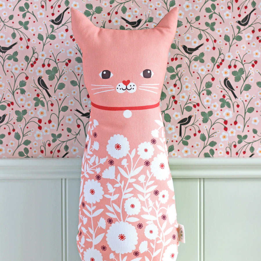 A pink cat-shaped decorative pillow with a floral pattern, featuring a simple embroidered face, set against a floral wallpaper background.