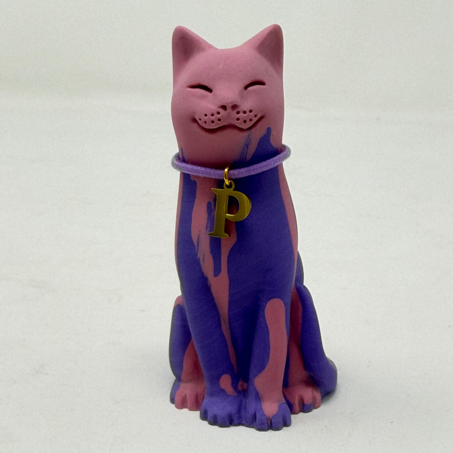 A purple and pink lucky cat-shaped resin figurine wearing a purple collar with a gold letter 'P' charm