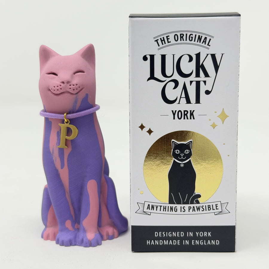 A pink and purple Lucky Cat wearing a purple collar with a gold letter 'P' charm, standing next to its white, black, and gold retail box 'The Original Lucky Cat York'.