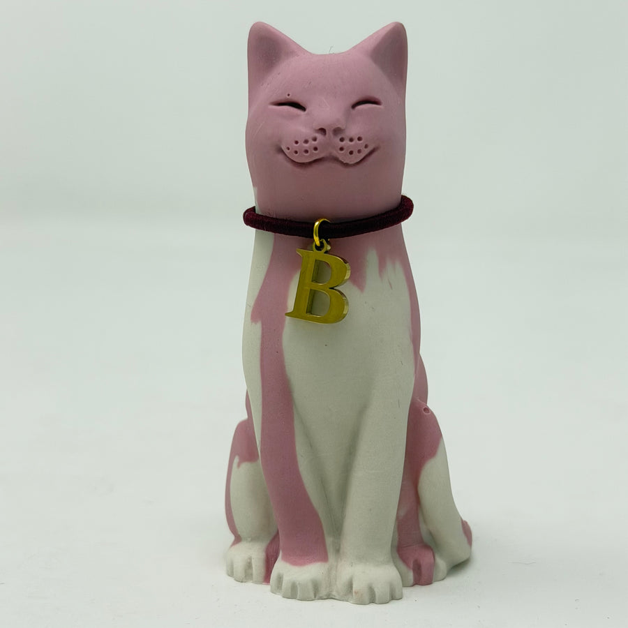 A pink and white lucky cat resin figurine wearing a dark red collar with a gold letter 'B' charm