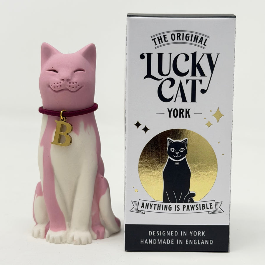 A pink and white resin Lucky Cat figurine wearing a burgundy collar with a gold letter 'B' charm, standing next to its branded box "The Original Lucky Cat York"