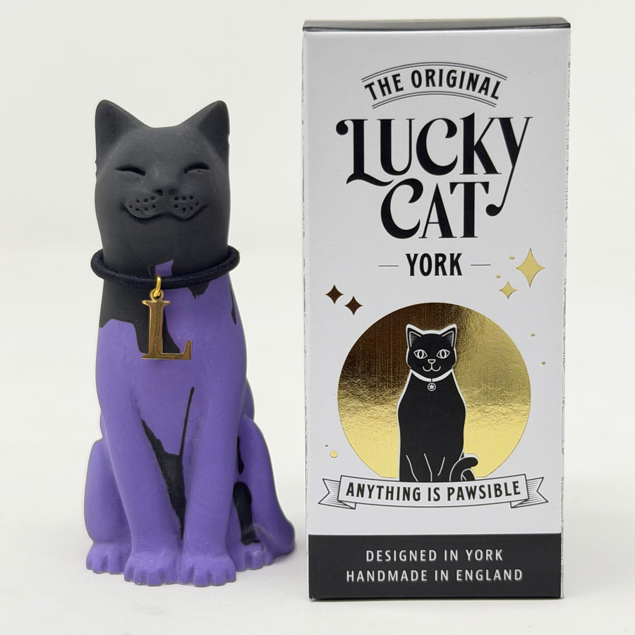 A purple and black cat figurine wearing a gold collar with a letter 'L' pendant, standing next to its white, black, and gold branded packaging box labeled 'The Original Lucky Cat York'.