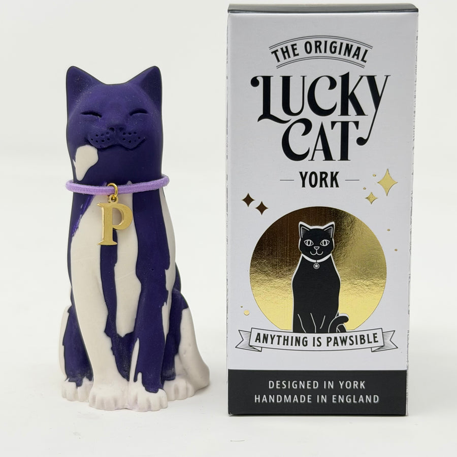 A purple and white resin Lucky Cat wearing a lavender collar with a gold letter P charm, shown next to its white, black, and gold branded box 'The Original Lucky Cat York'.