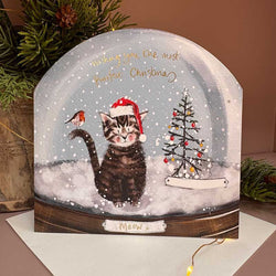 A decorative Christmas card shaped like a snow globe, featuring a tabby kitten wearing a Santa hat in the snow, a small decorated tree, and gold script text