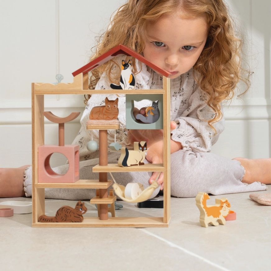 Child playing with a wooden toy house and cat figures on a light-colored floor.