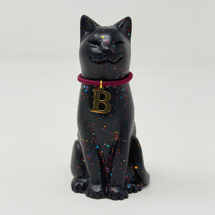 A black lucky cat figurine with multicolor glitter speckles, wearing a dark red collar with a gold initial 'B' pendant