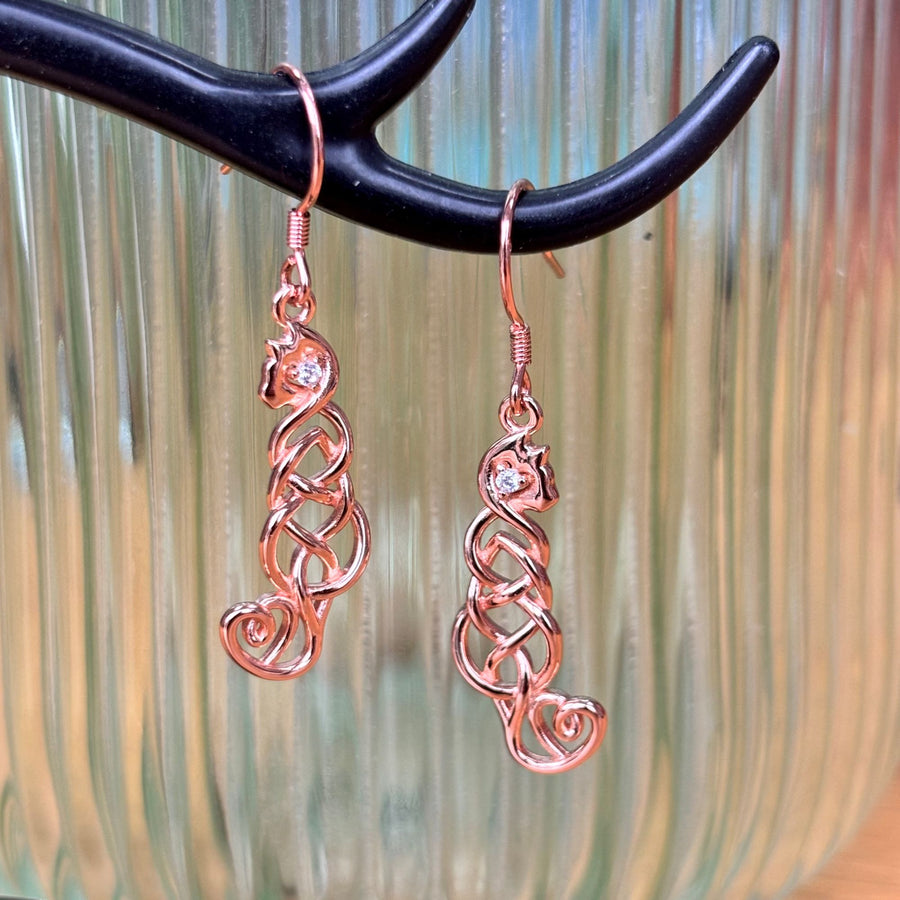 A pair of rose gold cat earrings with heart-shaped crystals, displayed on a black branch-like stand