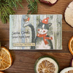 A rectangular bar of Santa Claws Spiced Apple Soap is displayed on a rustic wooden surface surrounded by dried orange slices, a green pine branch, and other botanical props.