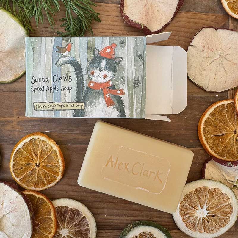 A bar of Alex Clark Speed Apple Soap shown next to its illustrated packaging box on a rustic wooden surface surrounded by dried citrus slices and greenery.