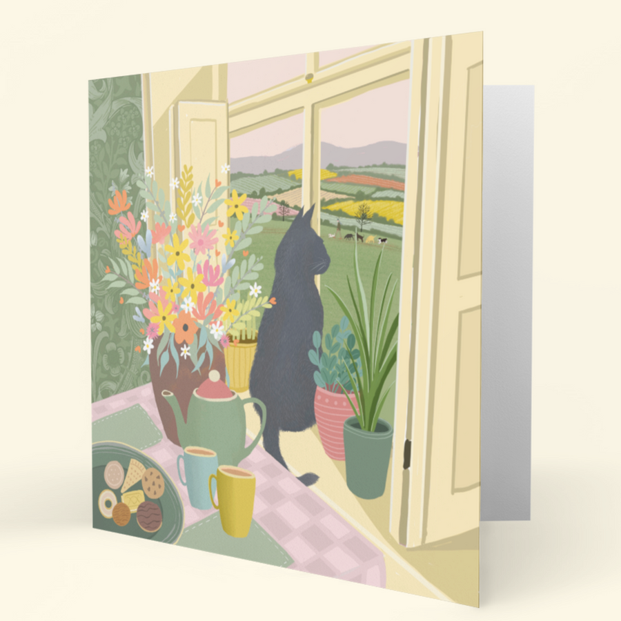 A folded greeting card featuring a black cat sitting by an open window, looking out over a colourful countryside landscape