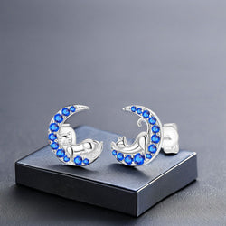 Silver moon-shaped earrings with blue stones and sleeping silver cats on a grey background