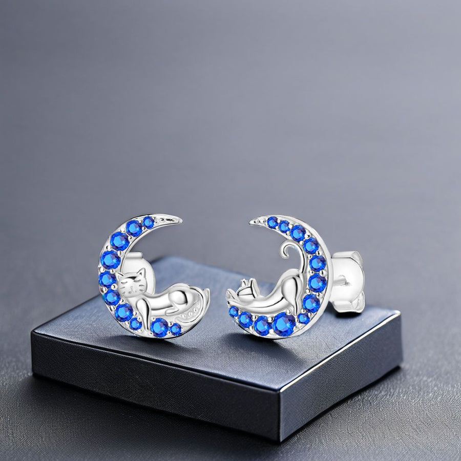 Silver moon-shaped earrings with blue stones and sleeping silver cats on a grey background