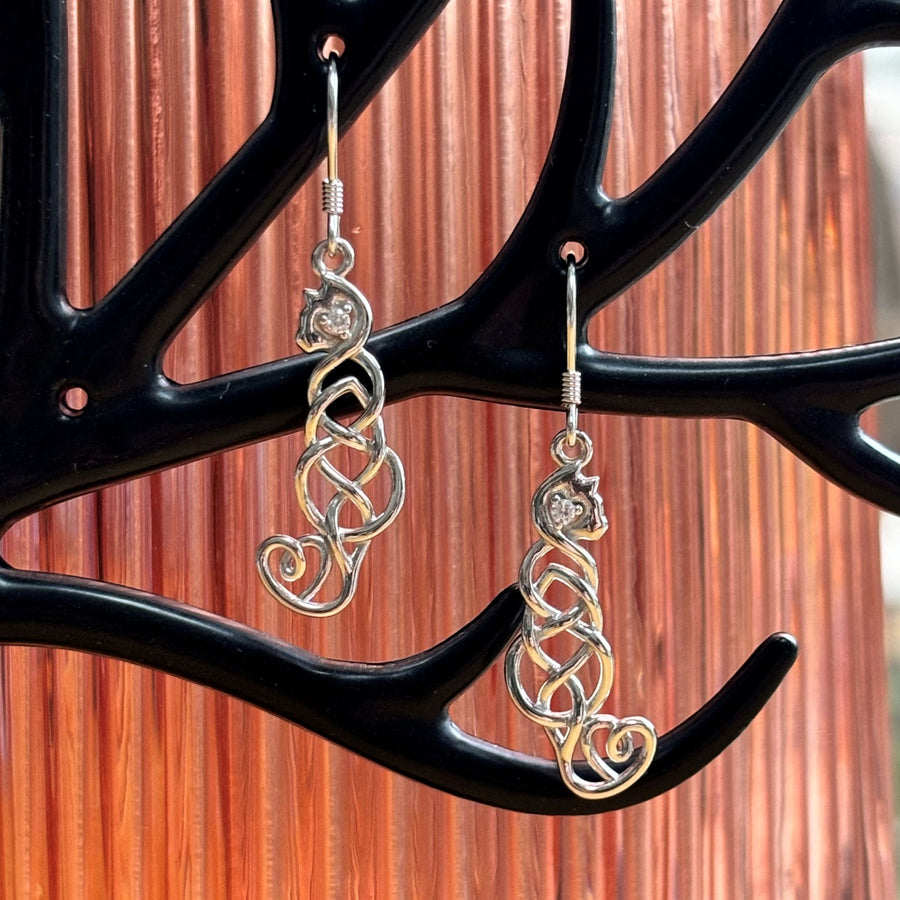 A pair of sterling silver cat earrings with heart-shaped crystals, displayed on a black branch-like stand