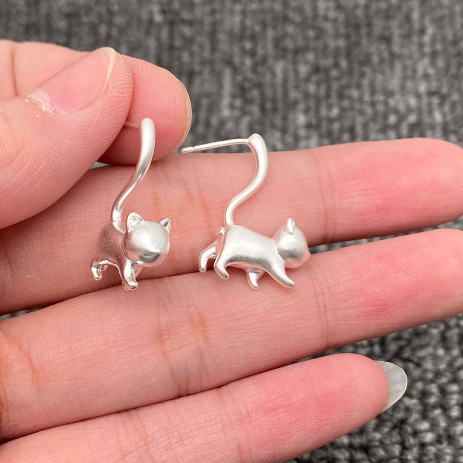 Silver cat-shaped earrings held between fingers on a textured surface